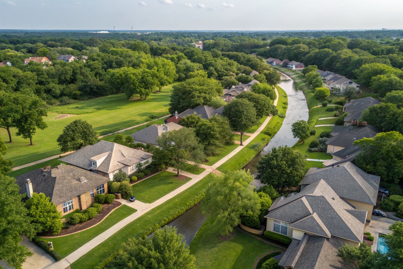 Aerial view of a suburban neighborhood of shingle-roofed homes beside a winding, tree-lined creek