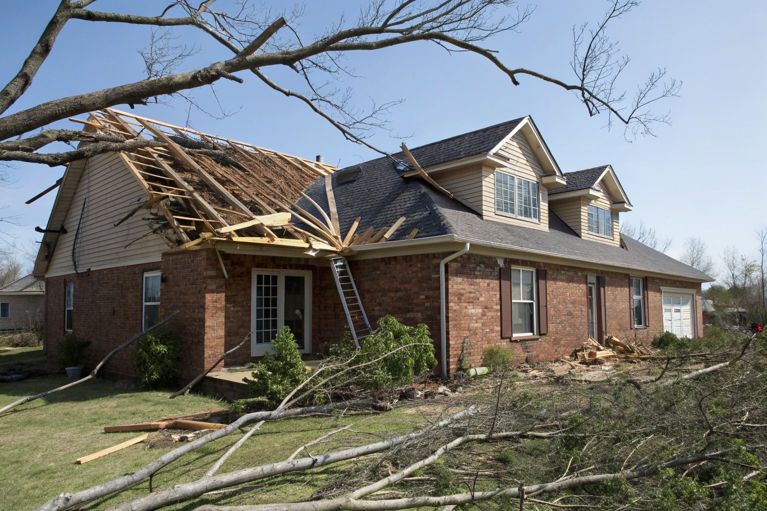 A brick house with part of its roof torn open by wind, rafters showing and branches across the yard